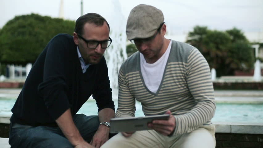Male friends with tablet sitting by the fountain, slow motion
