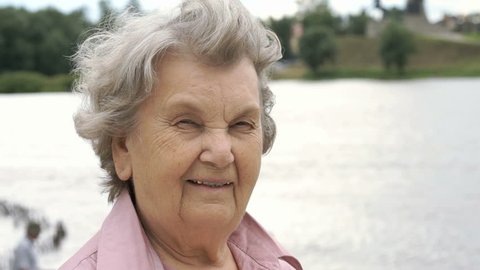 Portrait of smiling mature old woman with gray hair aged 80s on the background of river in summer. Slow Motion