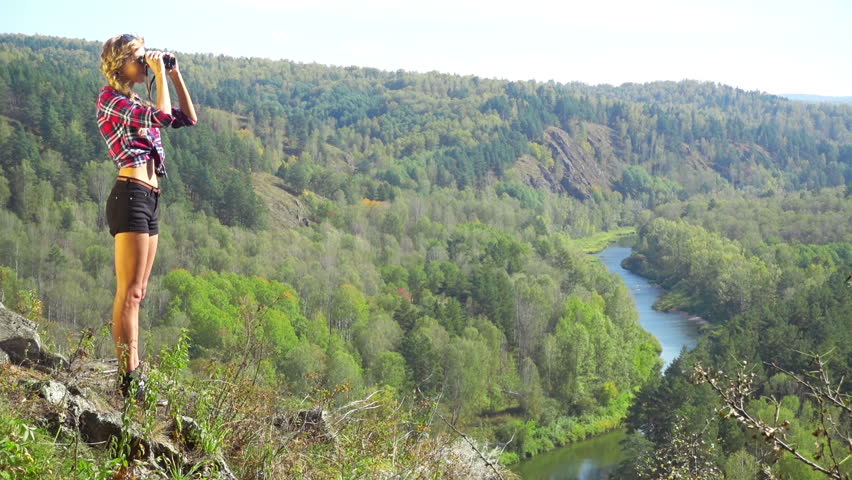 Young blonde woman tourist on a cliff looking through binoculars on the landscape with the river. Russia, Siberia, Salair
