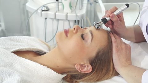 Cosmetologist applying iontophoresis on client's face. Female hands moving ball head of machine along girl's forehead. Pretty caucasian woman getting physiotherapeutic treatment for her facial skin