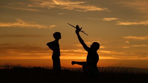 A happy family. The boy on his father's shoulders is playing with an airplane toy. Silhouette at sunset by the lake. at this time a real airplane is flying in the sky