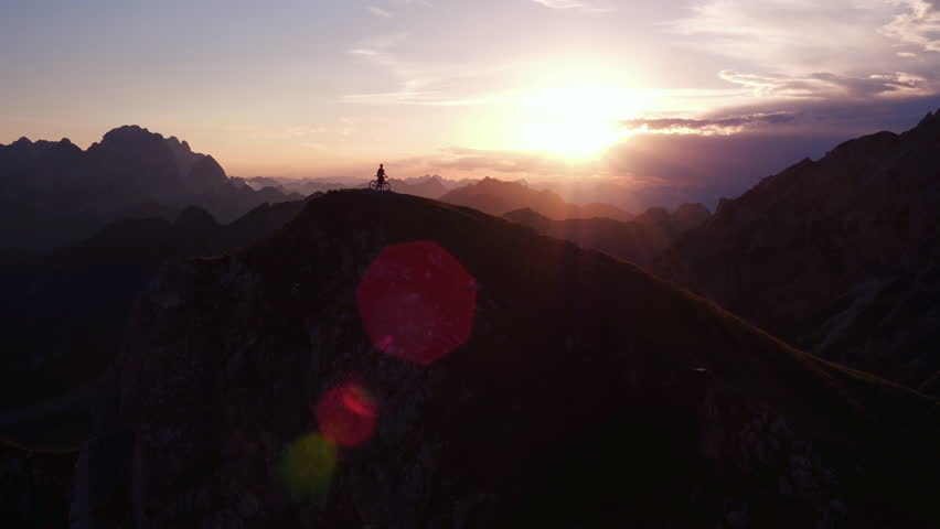 Aerial - Flyby adult mountain biker standing with his bike on top of the mountain at sunset