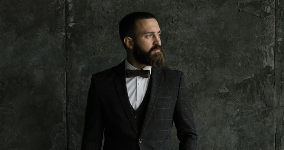 A bearded man ian a suit walk and looks out the window in a contemplative way. Medium shot