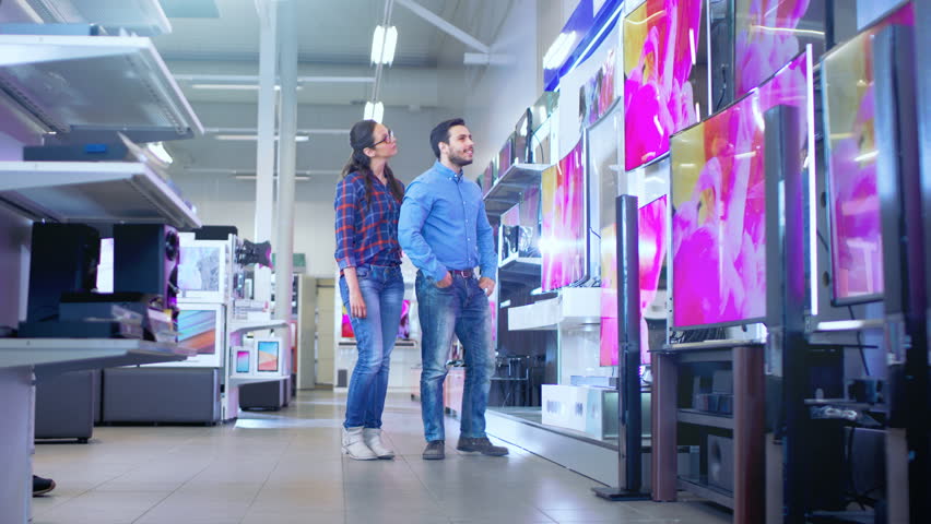 In the Electronics Store Professional Consultant Approaches Young Couple with His Expert Advice on Which of New Models of 4K TV's is Best for Their Home and Budget. Shot on RED EPIC-W 8K Helium Camera Royalty-Free Stock Footage #30542221