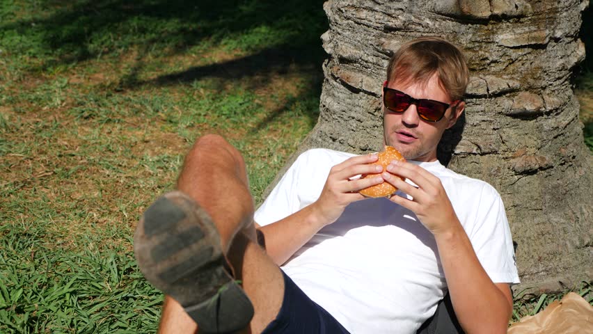 Handsome man sitting on the grass in the park under a tree, reading book, 4k, slow-motion