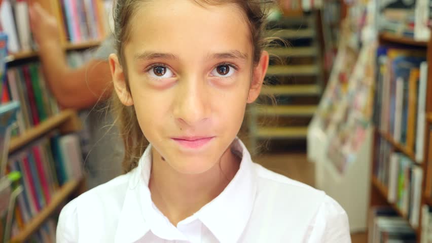 Portrait of a girl 8 - 12 years old, standing in the library. Bookshelves of a bookcase in the background. 4k, slow motion