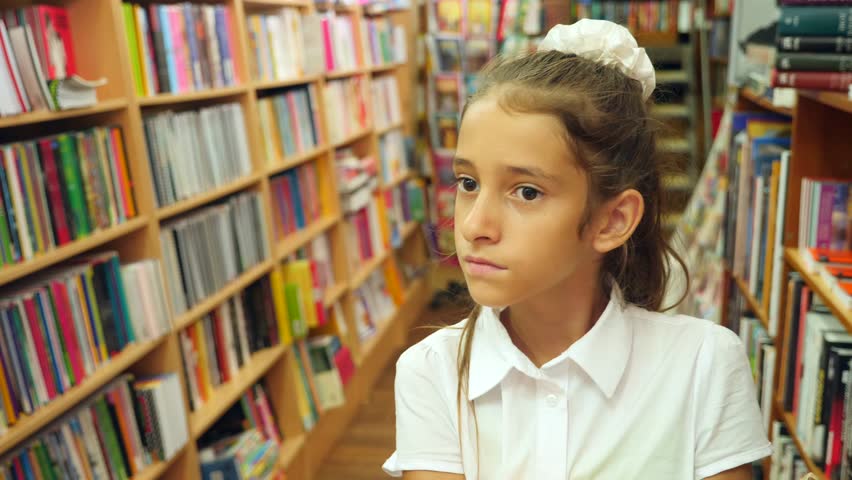 Portrait of a girl 8 - 12 years old, standing in the library. Bookshelves of a bookcase in the background. 4k, slow motion