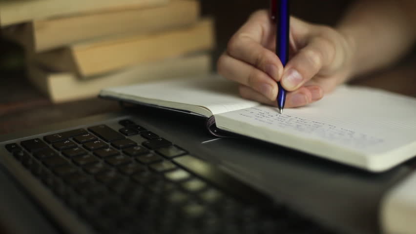 Woman hand writing in notebook (HD) laptop and a stack of books blur in the