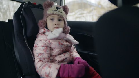 Little Girl in Winter Clothes Eats in an Baby Car Seat. a Winter Trip in the Car, the Child Looks Into the Camera and Shows His Displeasure. She is Dressed in a Winter Jacket and a Hat.