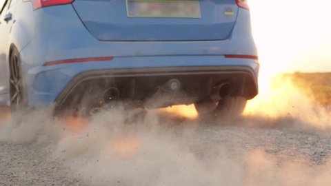 SLOW MOTION CLOSE UP: Sports car starts and quickly drives away from a dusty dirt road raising sand while performing burnout against beautiful golden summer sunset. Sportive auto scorching the tires