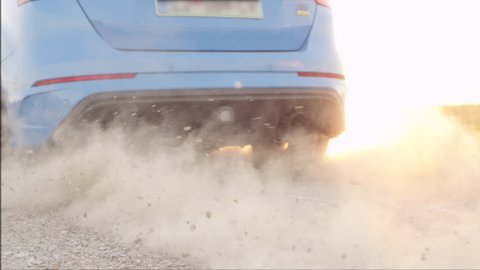 SLOW MOTION CLOSE UP: Sports car starts & quickly drives away from a dusty macadam dirt road raising sand, rocks and stones at dusk. Sportive auto performing burnout at beautiful golden summer sunset