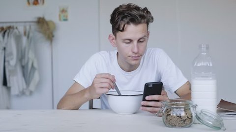 Bored and unimpressed millennial hipster internet user scrolls through smartphone applications, while slowly eats his organic breakfast with bran flakes and almond milk