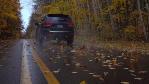 SLOW MOTION CLOSEUP: Black SUV car driving along an empty forest road, over fallen autumn tree leaves in reainy fall. Black jeep car driving fast along the wet slippery road, swirling colorful leaves