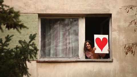 Young adult woman shows to the window a hand painted red heart and to her Valentine. Eastern Europe, Bucharest, Romania.
