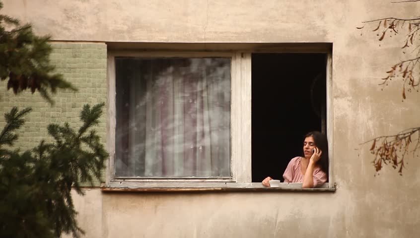 Young adult woman drinks coffee and speaks on mobile phone to the window. Eastern Europe, Bucharest, Romania.
