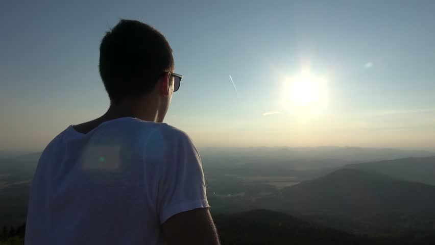 Young man rest on railing and looking into the sun - view of the countryside from hill / high place. Landscape in the background. Nice sunny day. Clear blue sky. 