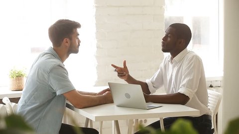 African american and caucasian young businessmen negotiating sitting at office desk with laptop, two satisfied partners close successful business deal with investor, handshake after making agreement