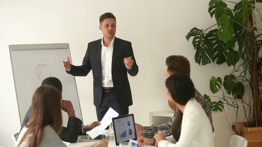 Successful caucasian businessman gives presentation in boardroom for multi-ethnic partners group, business coach answers questions, explains pointing on chart on whiteboard, interacting with audience - Powered by Shutterstock - Get 15% off with code: PIKWIZARD15