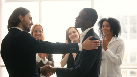 Boss promoting rewarding african american male worker, appreciating for good job, businessmen wearing suit congratulating shaking hands with applauding staff standing in office, employee recognition