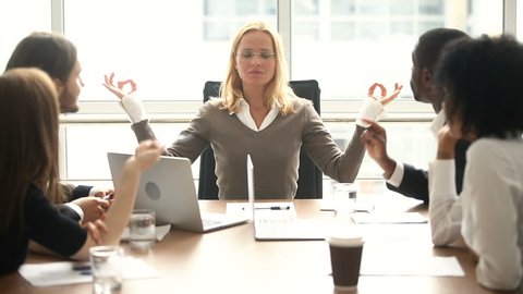 Calm young businesswoman meditating with eyes closed at meeting with multiracial colleagues, female boss keeping mental balance at stressful job, controlling emotional health, no stress at work, om