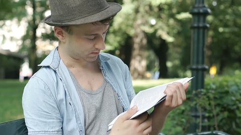 Young man finishes reading book in the park and relaxing, steadycam shot
