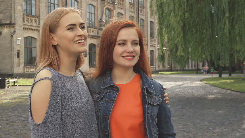 Women smiling and hugging. They look aside, then at each other and at camera. Girls standing outdoors near university
