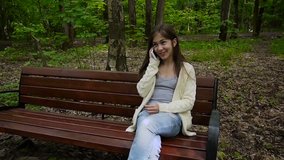 Young attractive girl speak to phone on bench. Summer park. Smile. HD video footage shooting static camera.