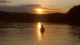 young girl stands in water and looks at sunset