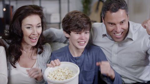 Happy caucasian family get very excited watching sports on television and the popcorn spills everywhere. In slow motion.