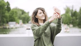 Happy young woman wearing a green sweatshirt is standing on a bridge and taking a selfie. She is using her smartphone and having a good time. Left to right pan real time establishing shot
