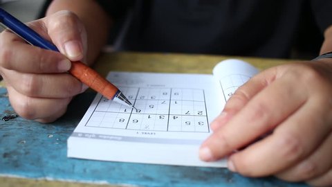 Female hands trying to solving Sudoku puzzle on books with pencil of blue wooden office desk. Originally called Number Place.