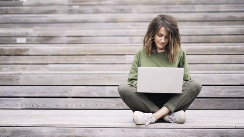 Crosslegged beautiful young woman wearing a green sweatshirt sitting on the stairs outside and typing at her laptop. Locked down real time establishing shot