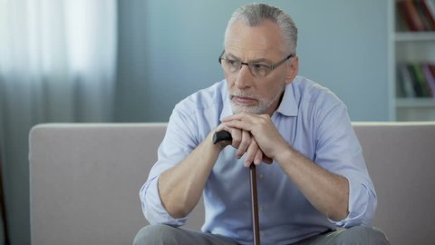 Ill man in his 50s sitting on couch, putting hands on walking stick. Depression