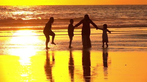 SLOW MOTION, CLOSE UP: Silhouetted Muslim family holding hands standing on dreamy sandy Kuta Beach in Bali watching splashing ocean at golden light sunset. Arab mother and children watching sea waves