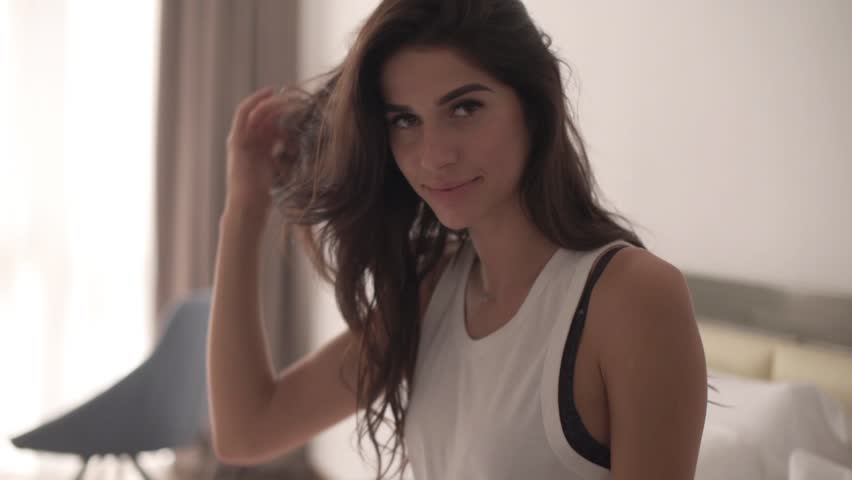 girl with dark hair sitting on the bed in the morning, cute smiles into the camera. White bedroom, Sun behind the window