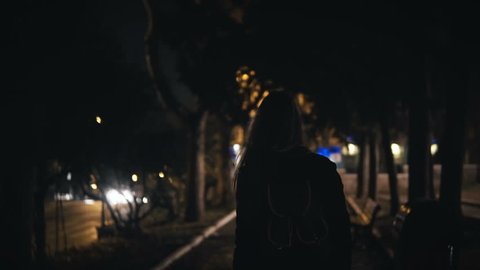 Back view of tourist woman with backpack walking through the dark park near the road late at night alone.