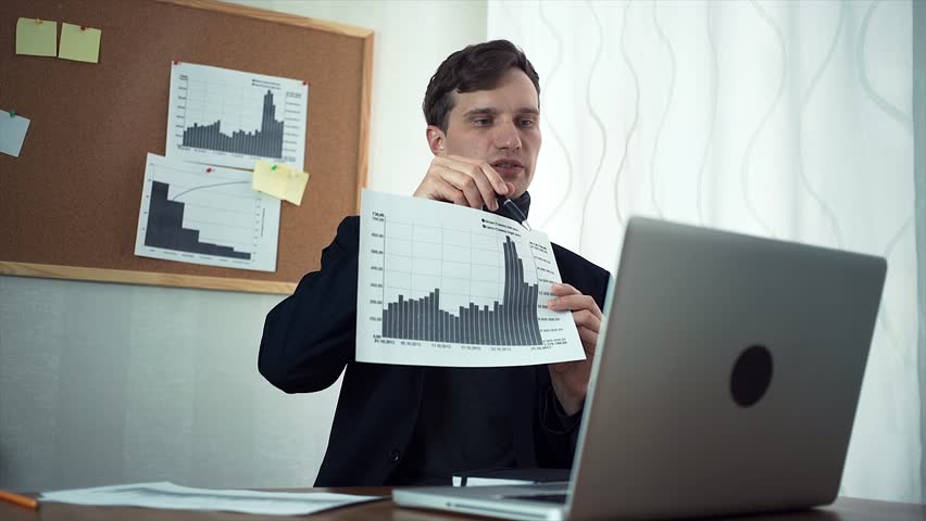 Young financial analyst chatting with client using laptop computer demonstrating printed data and analyzing graphs. A board with sticky notes and graphs on the wall behind Royalty-Free Stock Footage #30681259