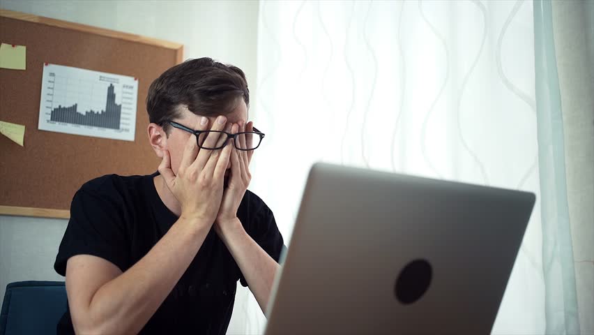 Tired young business man in the glasses rubs tired eyes. Freelancer working on the office using laptop. A board with stickers and graphs on the wall behind Royalty-Free Stock Footage #30681502