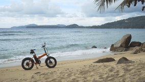 Battery powered bike stands on it's kickstand in the sand as gentle waves wash against this tropical beach paradise. UltraHD 4k video