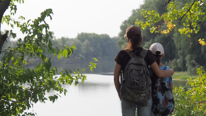 Tourists with backpacks look at the river. Mom is hugging her daughter on the river bank.
