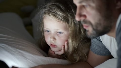 Tilt up of adorable little girl and her bearded father lying in bed and chatting while watching something on tablet in darkness