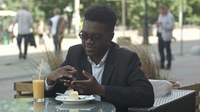 Handsome young afro american businessman using smart phone, messaging his girlfriend, eating at cafe