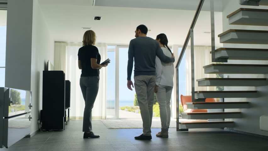 Professional Real Estate Agent Shows Stylish Modern House to a Beautiful Young Couple Who are in the Market for Purchasing/ Renting New Home. House Has Floor to Ceiling Windows and Seaside View.4K UHD