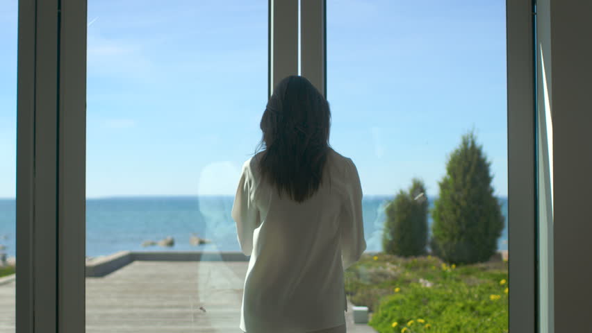 Following Shot of a Beautiful Brunette with Loose Hair Opening Doors of the Balcony and Walking onto Sunny Terrace with Seaside View. It's Cloudless Morning and She's Really Happy. 4K UHD. Royalty-Free Stock Footage #30700843