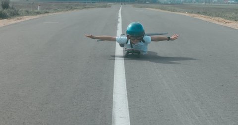 Little boy wearing helmet and styrofoam wings riding skateboard on a rural road, pretending to be a pilot. 4K UHD RAW edited footage