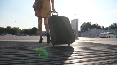 Business lady going to taxi parking from the airport with her luggage. Girl in heels stepping and roll suitcase on wheels. Woman walking with her suitcase along street. Travel concept. Slow motion