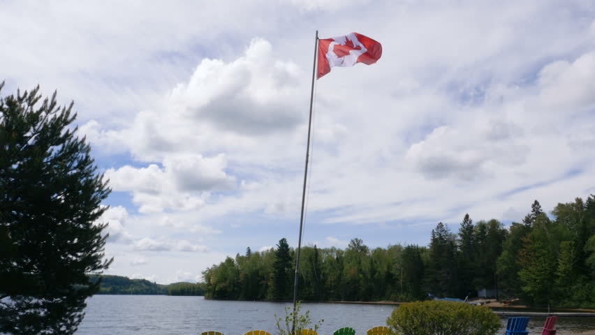 The Canadian flag flaps in the wind with Georgian Bay waters and islands in the background. Multicolored lounge chairs underneath. Long shot, low angle. 