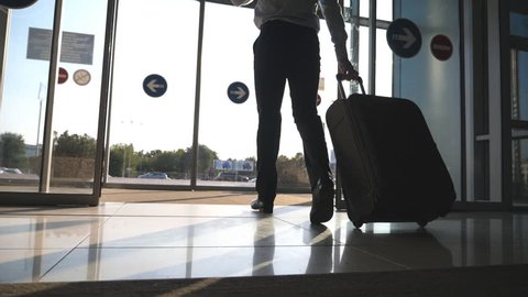 Man going through glass automatic door to the street and roll suitcase on wheels. Businessman walking from the airport with his luggage. Sun flare at background. Trip or travel concept. Slow motion