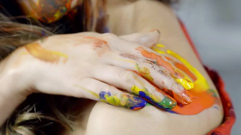 Young and glorious model is slowly painting her bare shoulder in studio with bright colors.