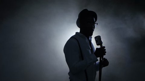 Man african american singer in the smoke and white light sings into a microphone and dance. Black background. Silhouette